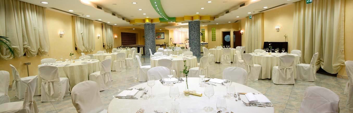 Uma sala de banquetes com mesas redondas preparadas para refeições, cadeiras brancas e um palco ao fundo.