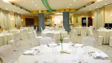 Uma sala de banquetes com mesas redondas preparadas para refeições, cadeiras brancas e um palco ao fundo.