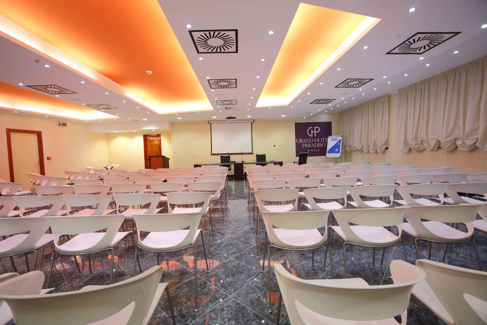 A conference room with rows of white chairs, a podium, a screen, and a banner reading "Grand Hotel Paradiso".