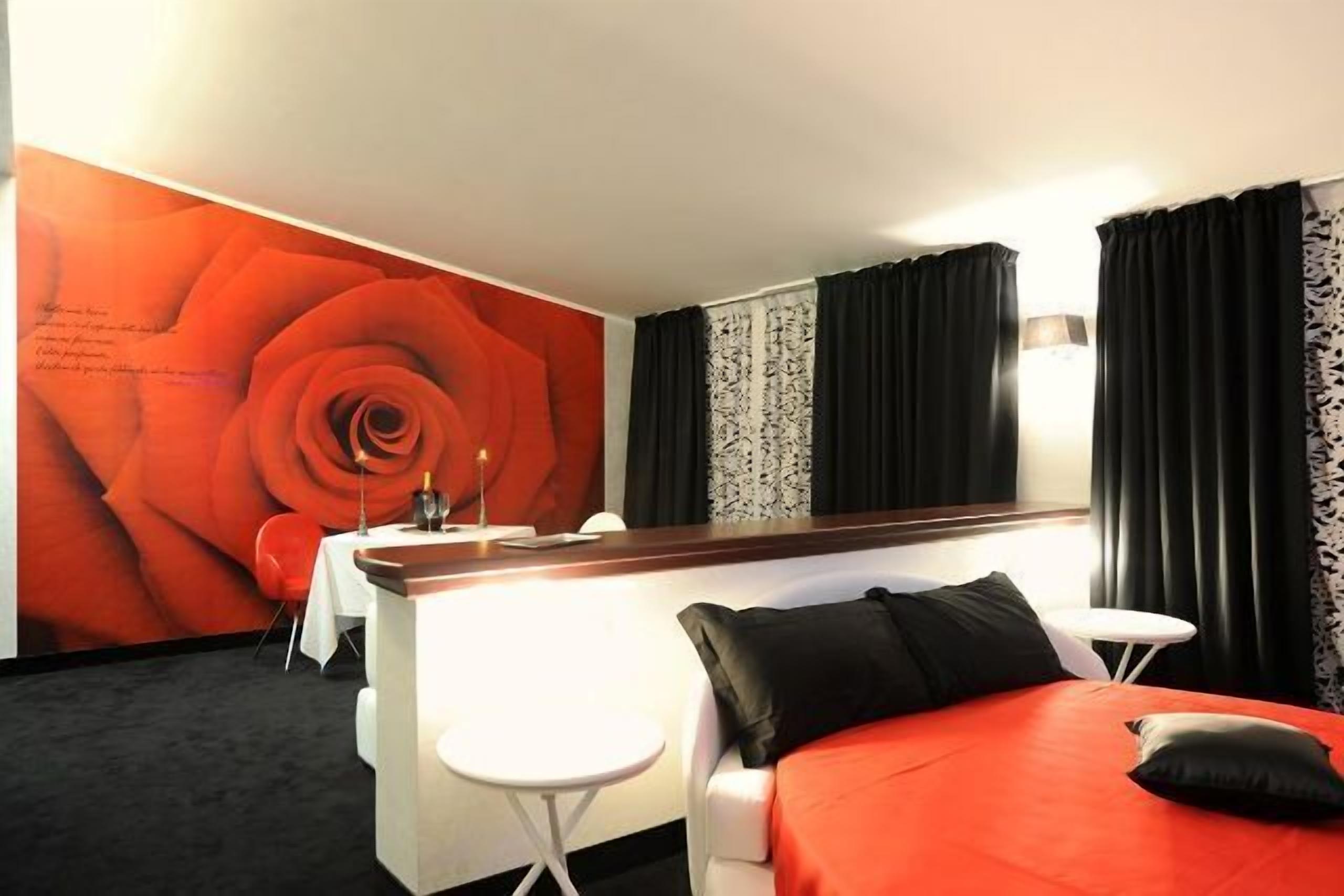 A hotel room with a large wall mural of a rose, a bed with red bedding, a desk with a chair, and a small table.
