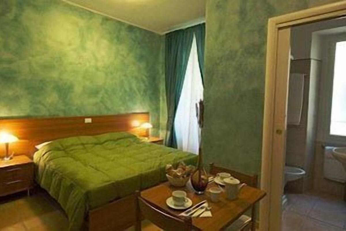 A hotel room with a bed, bedside lamps, a table with cups and a teapot, and a view of the bathroom.