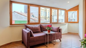 Una habitación de esquina con un sofá marrón, una mesita con una planta en maceta y ventanas amplias con vistas a edificios.