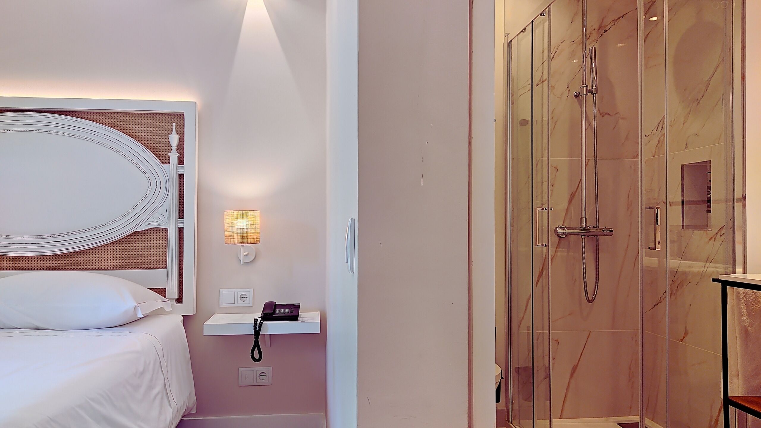 A hotel room with a bed, a bedside table with a lamp, a phone, and a glass-enclosed shower.