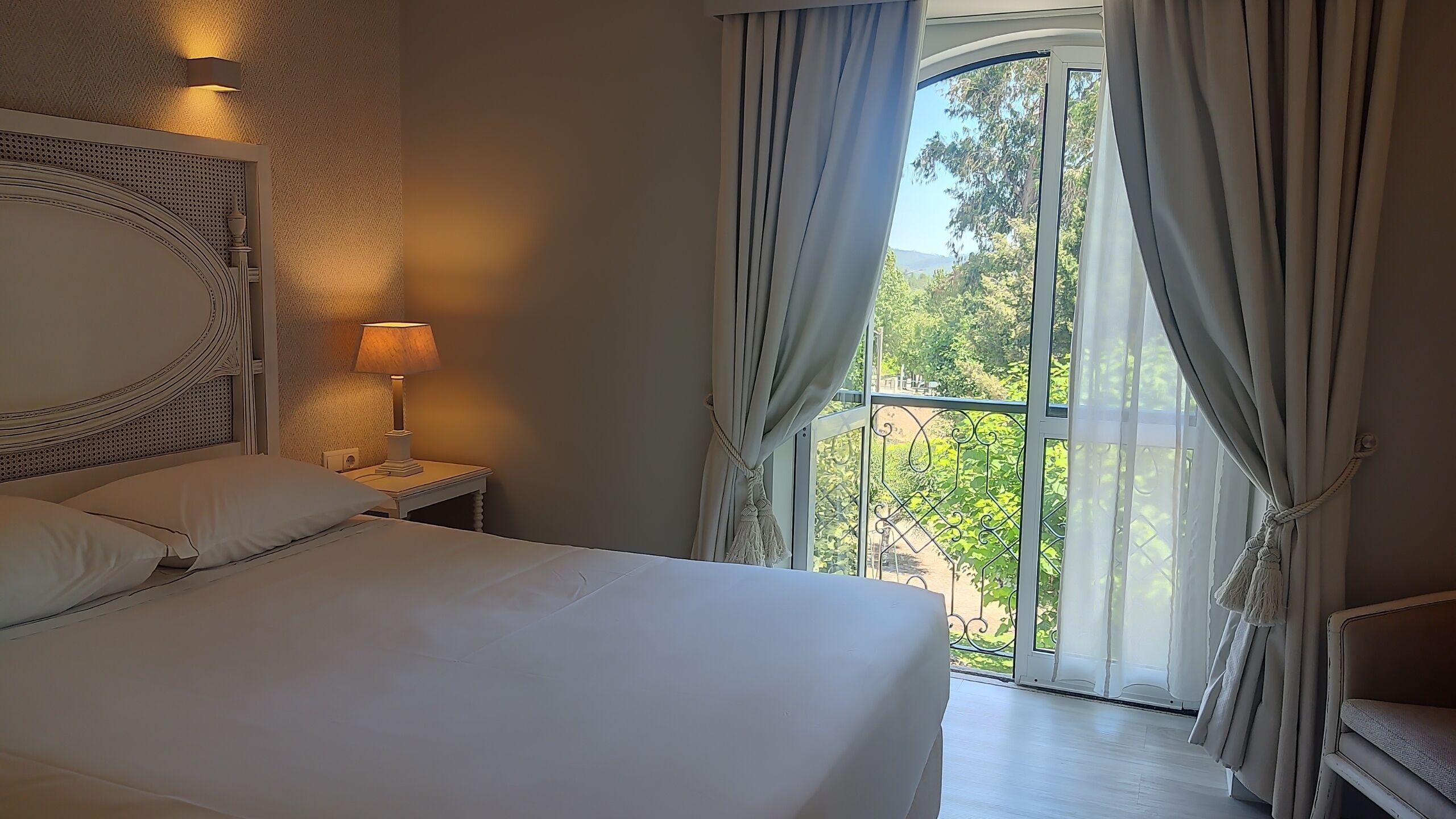 Quarto de hotel com uma cama grande, luminária, mesa de cabeceira e vista para o verde através de uma janela com cortinas.