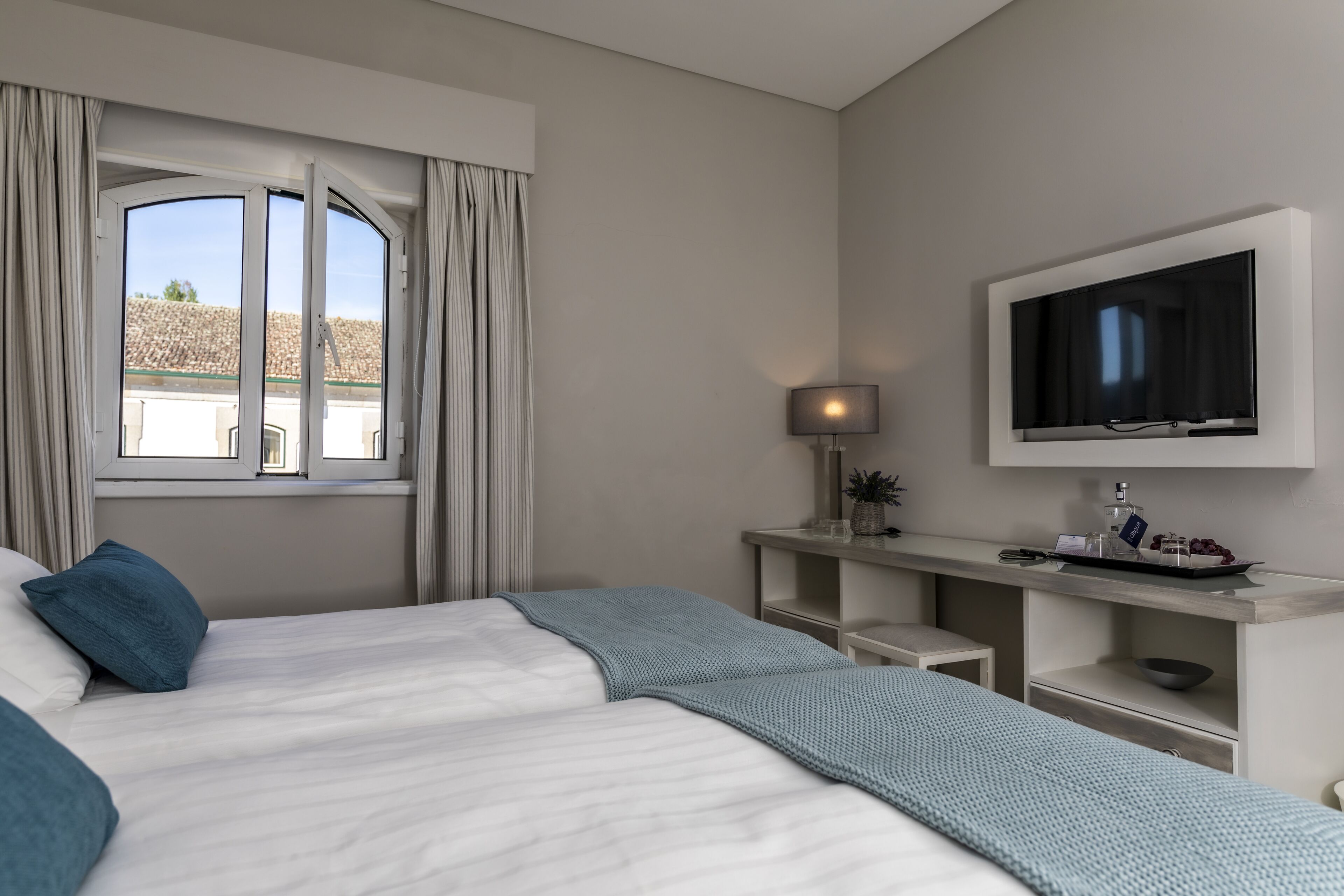 Una camera d'albergo con un letto, una TV a muro, una finestra con tende e un tavolino con una lampada e un vassoio di frutta.