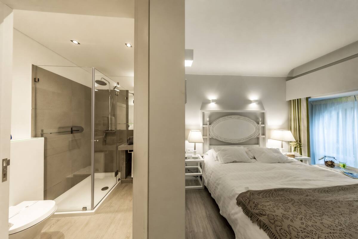 Une chambre d’hôtel moderne dotée d’une douche en verre, d’un grand lit agrémenté d’une tête de lit décorative, de lampes de chevet et offrant une vue sur l’extérieur grâce à une fenêtre munie de rideaux.