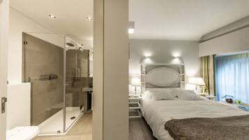 Habitación de hotel moderna con ducha de vidrio, una cama grande con cabecera decorativa, lámparas de noche y vistas al exterior a través de una ventana con cortinas.