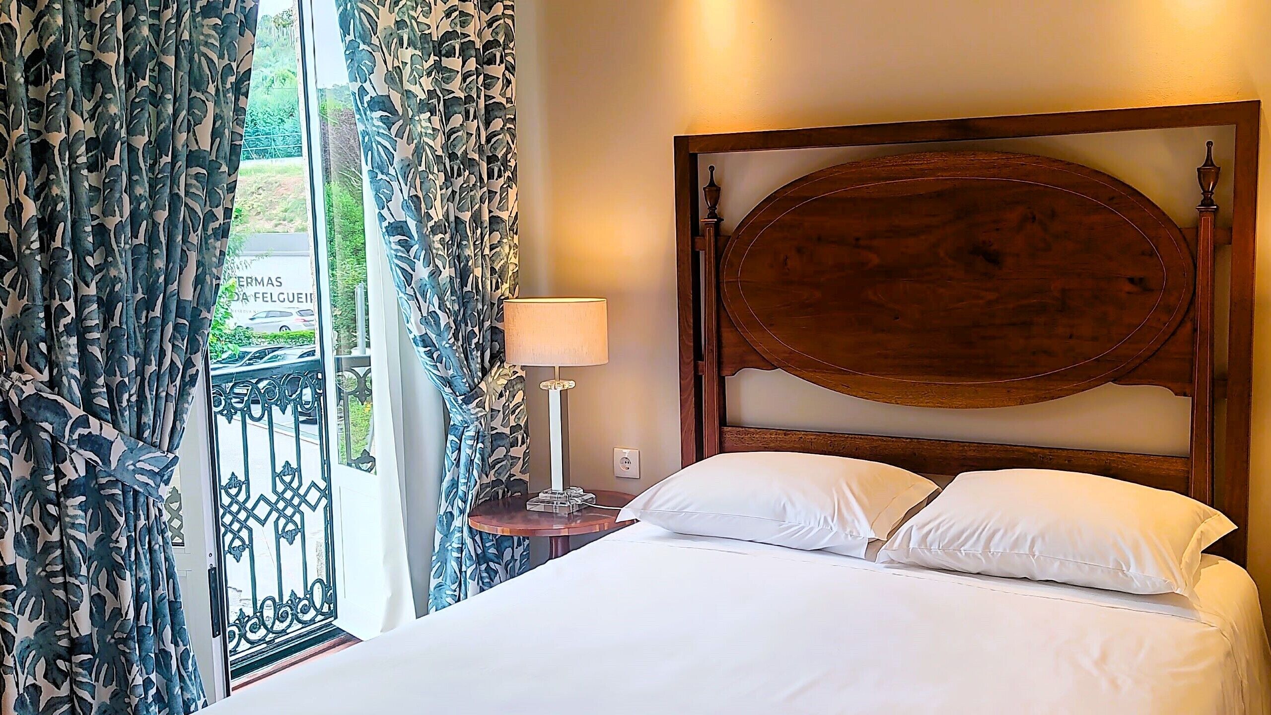 Een hotelkamer met een bed, nachtkastje, lamp en uitzicht op de omgeving door een raam met patroon gordijnen.