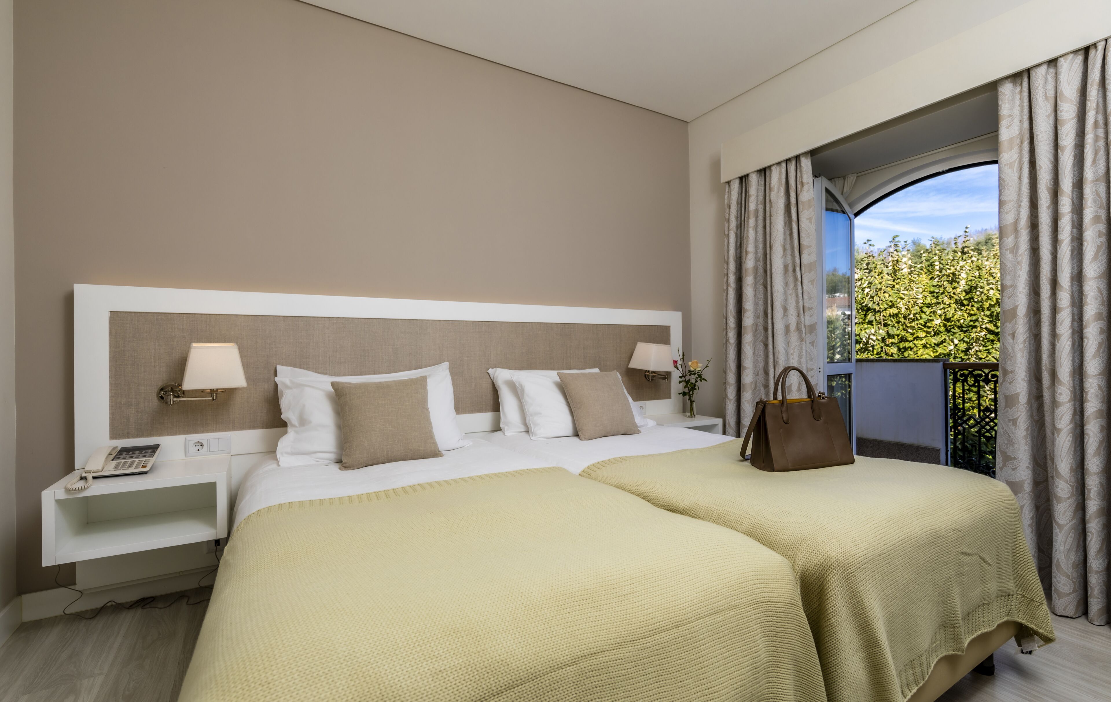 A hotel room with a large bed, two bedside tables, a nightstand, a lamp, a handbag, and a view of greenery through a window.