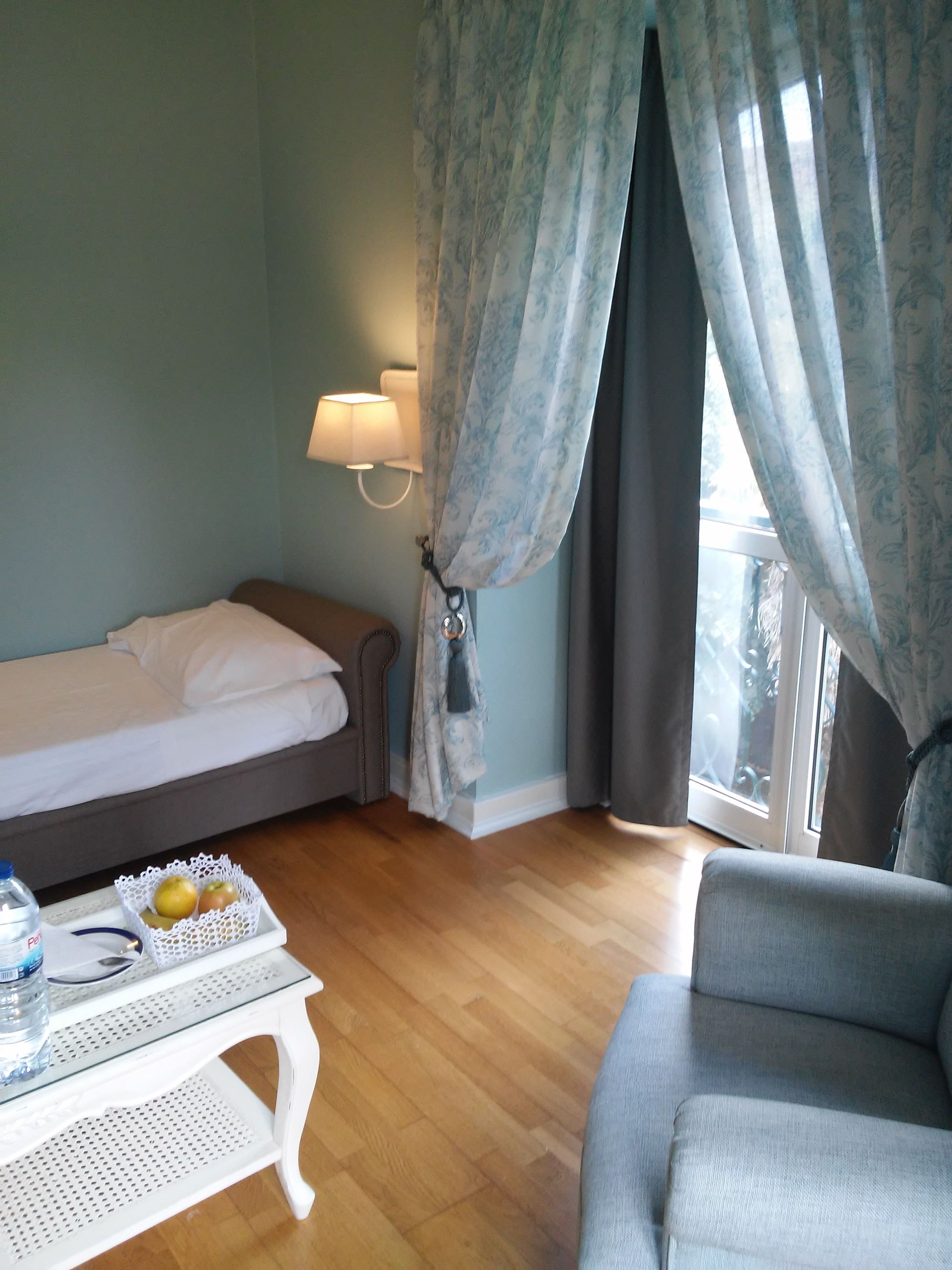 A hotel room with a bed, a small table with a water bottle and fruit, a chair, and a window with curtains.
