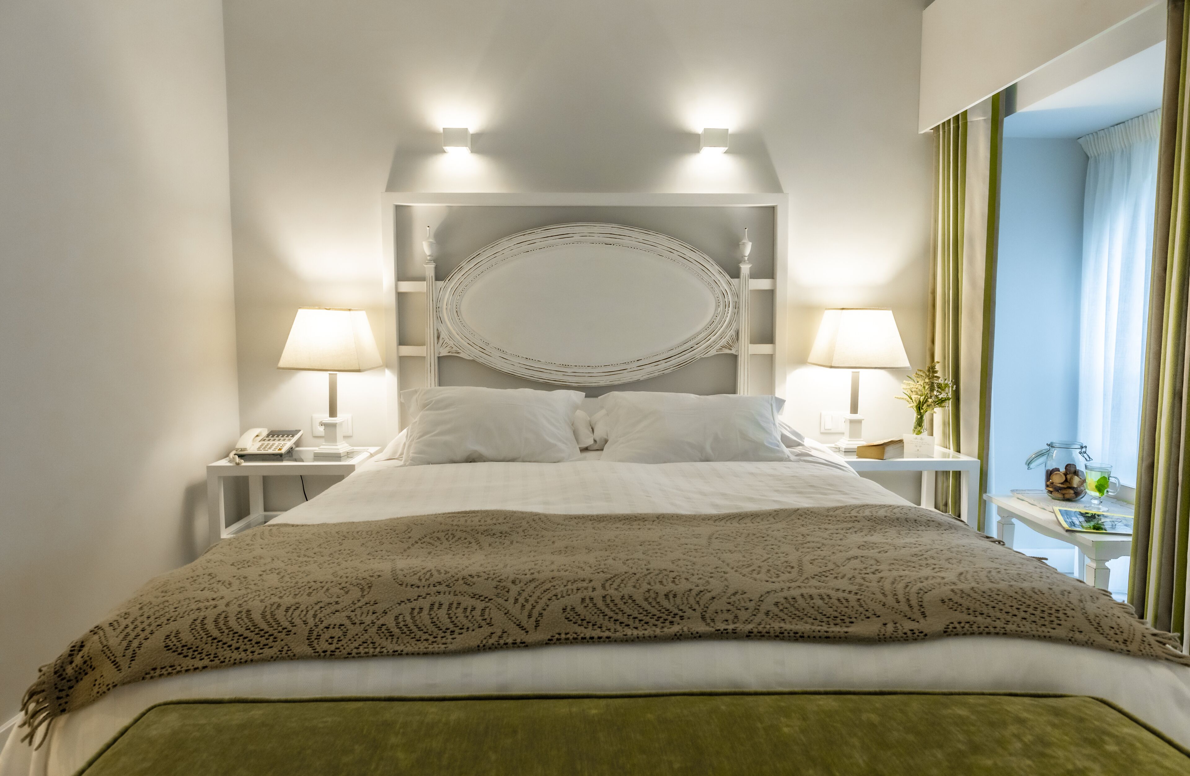 Una cama bien hecha con una colcha estampada, dos lámparas de noche, un teléfono junto a la cama y una mesita con un vaso y una planta.