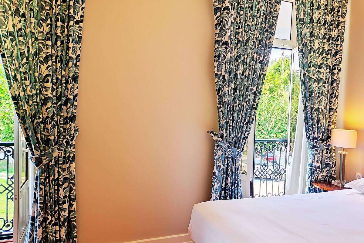 Une chambre d’h ôtel avec un lit, des rideaux, une fenêtre donnant sur un paysage verdoyant et une table de chevet avec une lampe.