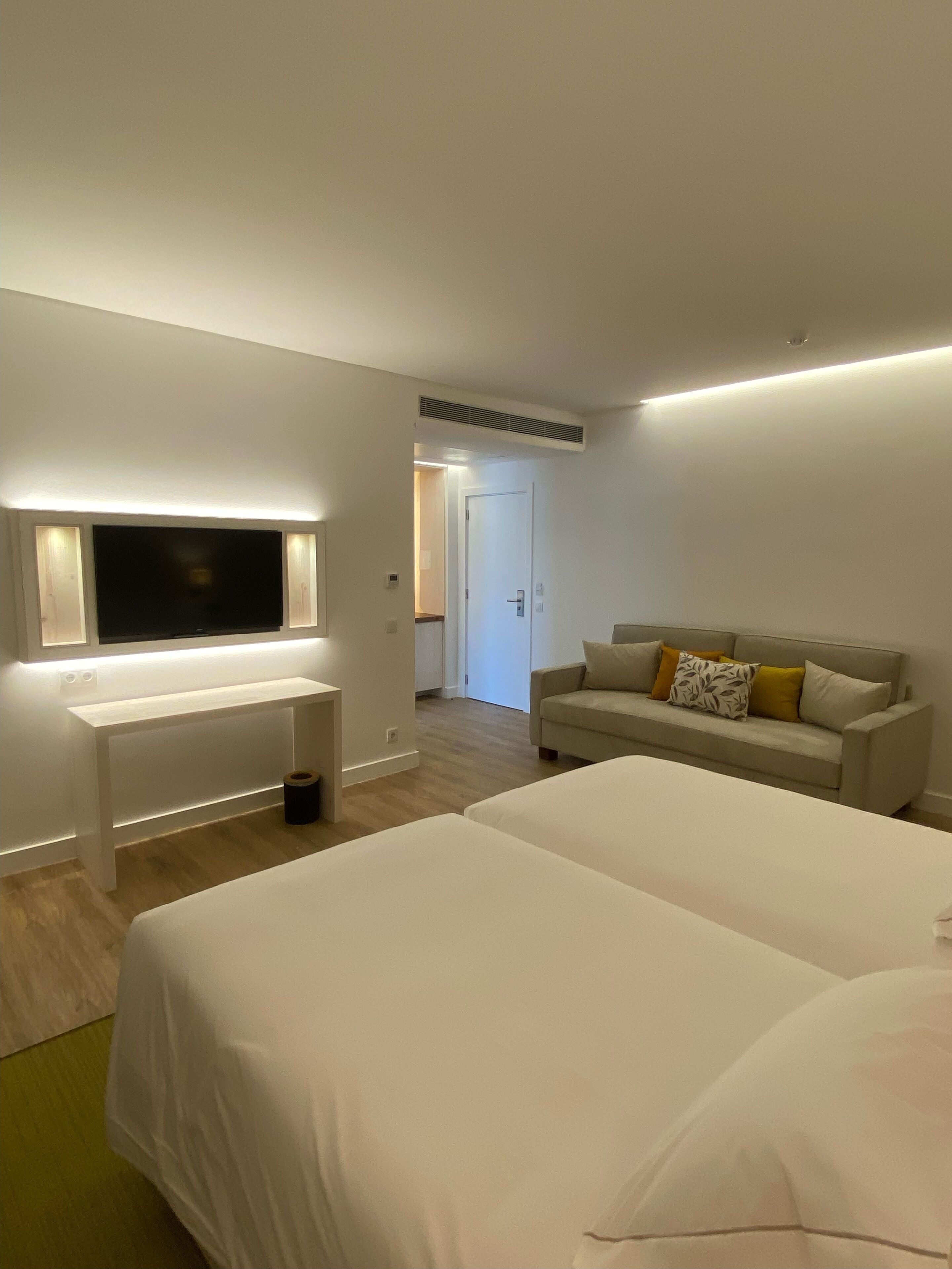 A hotel room with a large bed, a bedside table, a TV mounted on the wall, and a sofa with cushions.