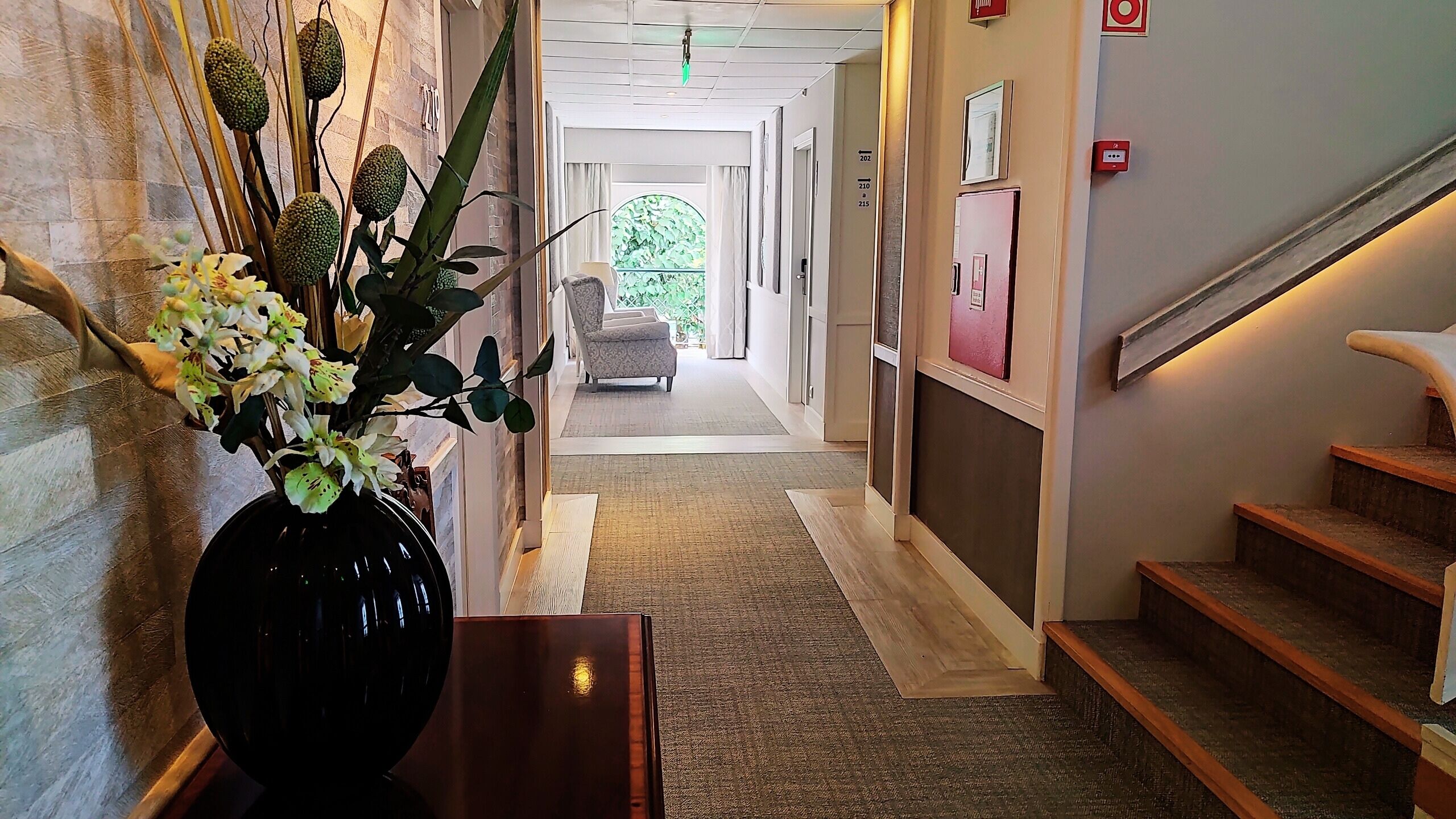 Um corredor de hotel com escadas de madeira, um vaso com flores e uma cadeira ao fundo.
