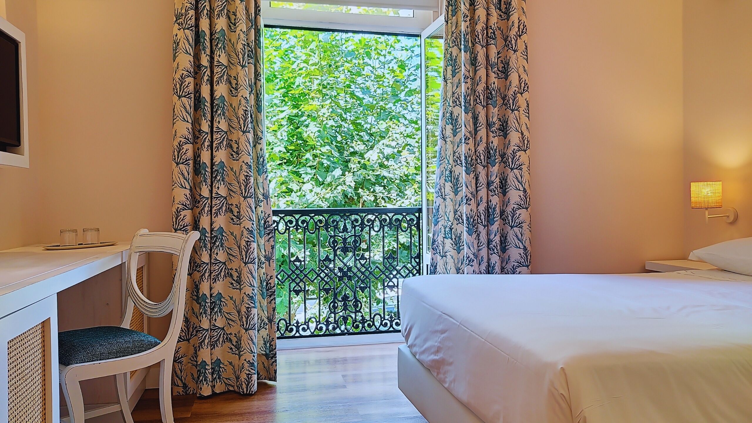 A bedroom with a bed, a desk, a chair, and a balcony with a view of greenery.
