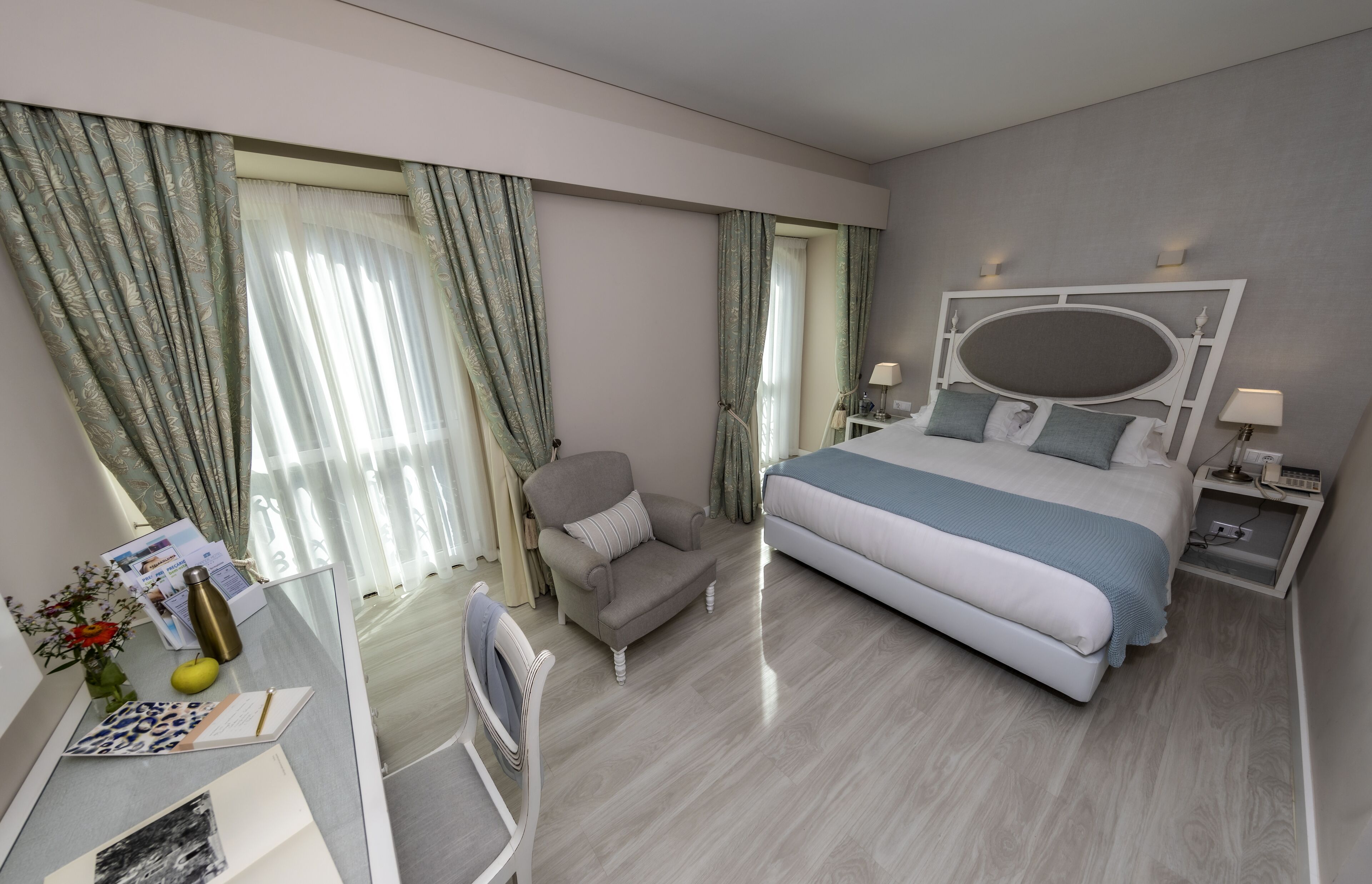 A hotel room with a large bed, a desk with a chair, a bedside table with a lamp, and a window with curtains.