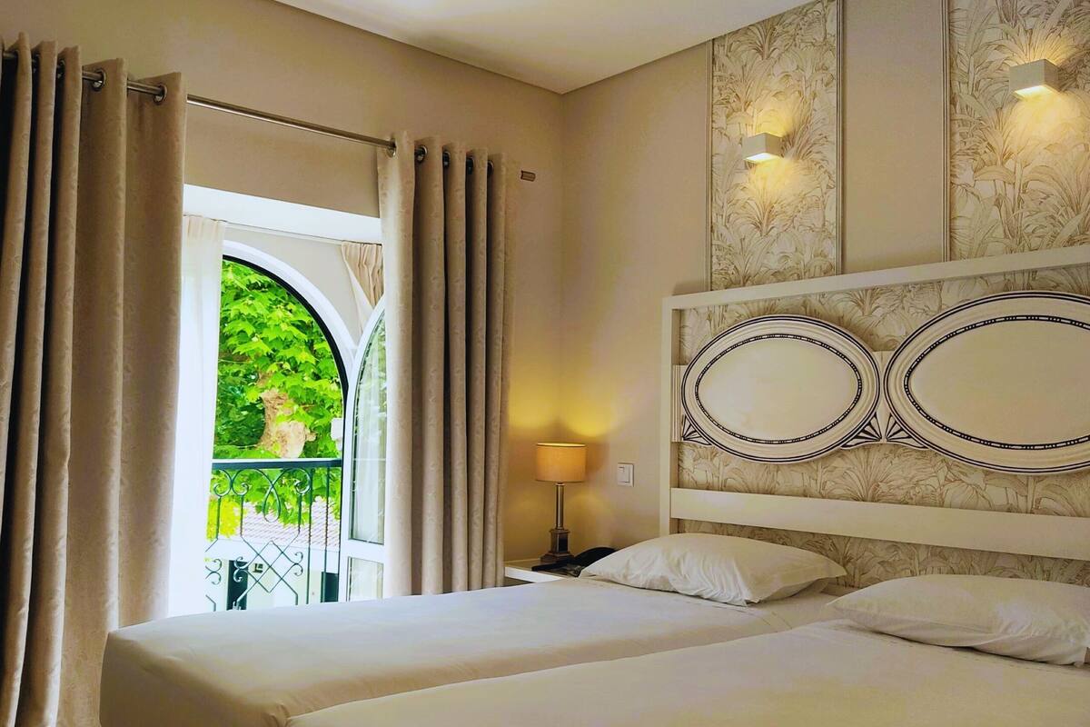 Une chambre d’hôtel avec deux lits, une fenêtre avec des rideaux, une lampe et un panneau décoratif au mur.