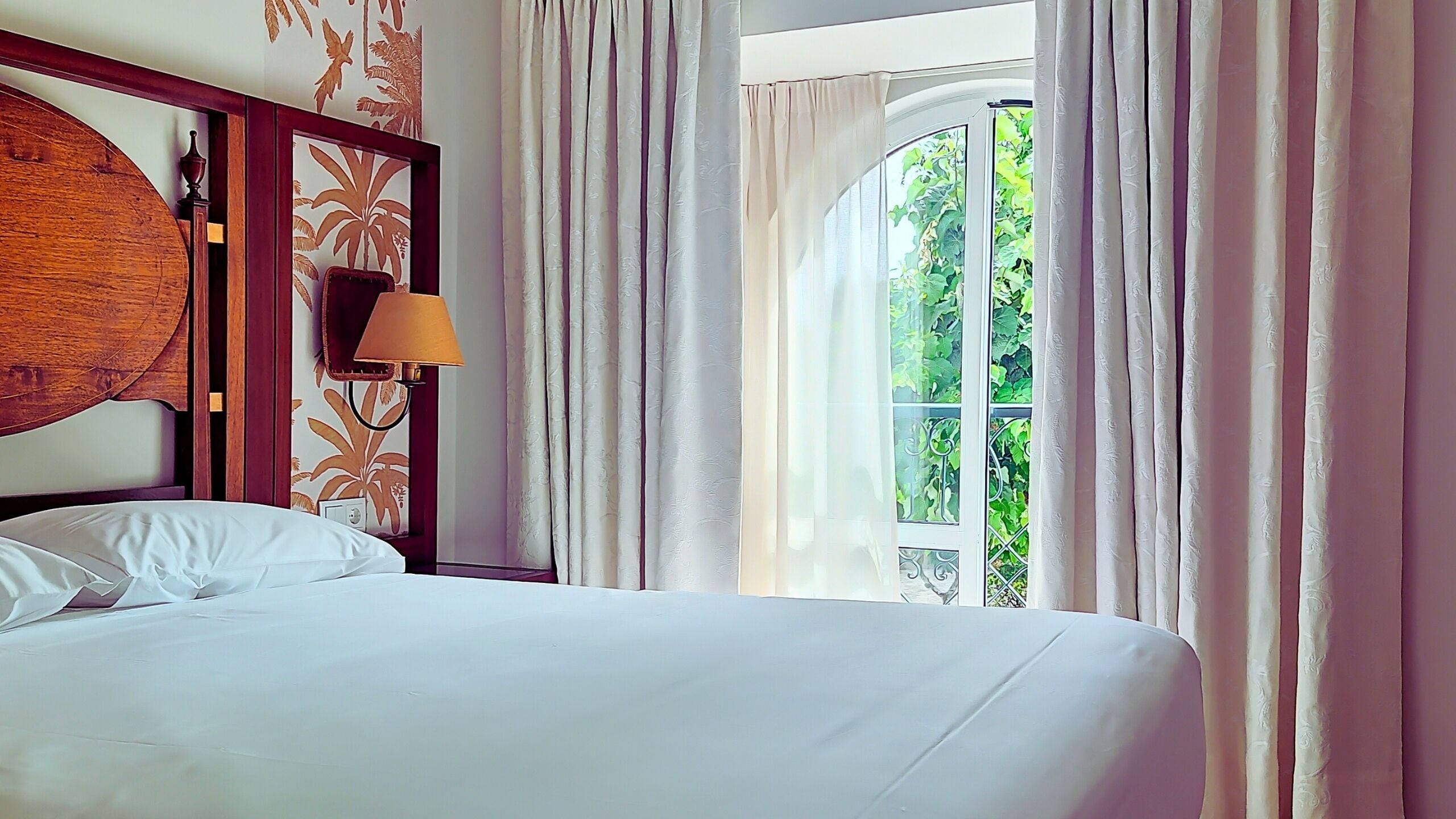 Ein Hotelzimmer mit einem großen Bett, einem Holzkopfende, einer Wandleuchte und einem Fenster mit Vorhängen.