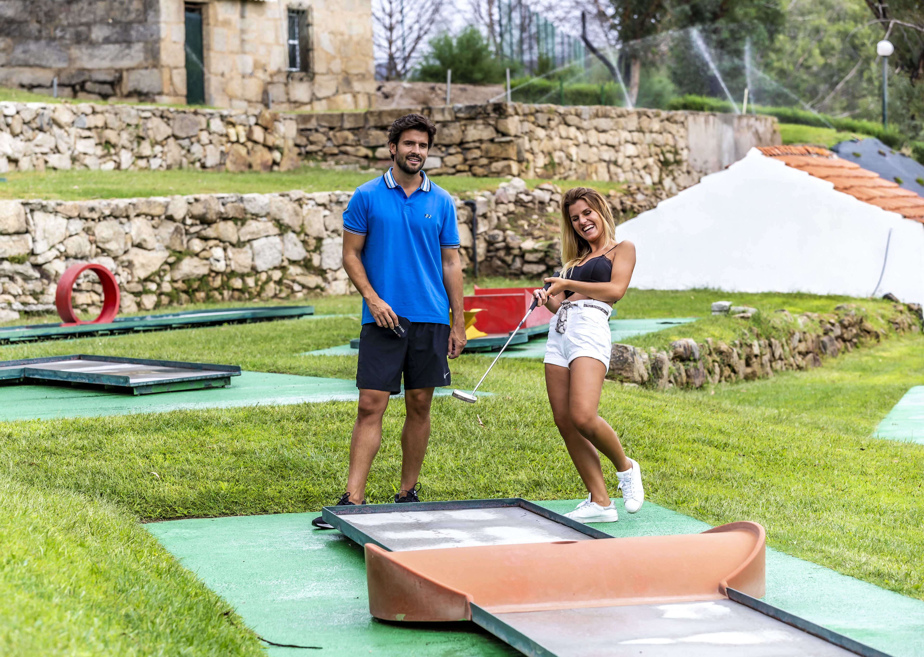 Un hombre y una mujer en un campo de golf, con un muro de piedra y una tienda de campaña al fondo.