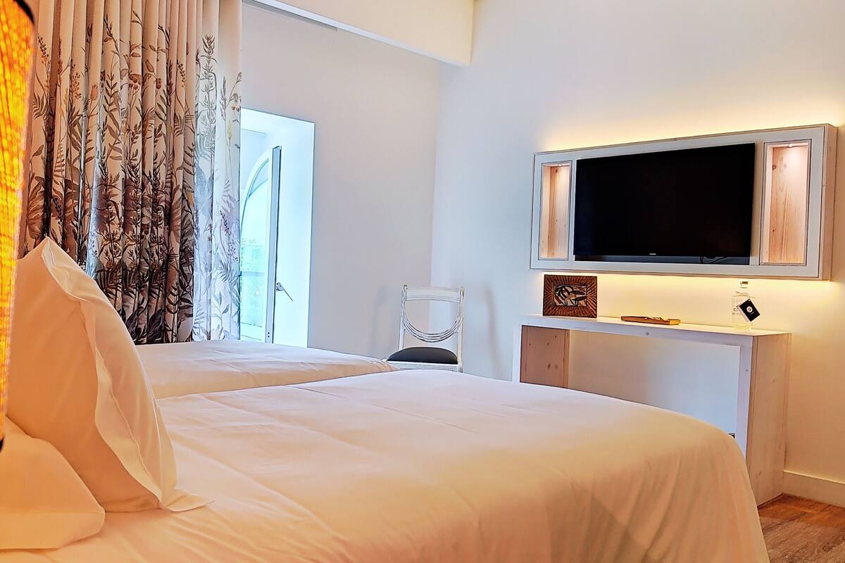 Une chambre d’hôtel avec deux lits, un téléviseur fixé sur une console blanche, une chaise et une fenêtre avec des rideaux.