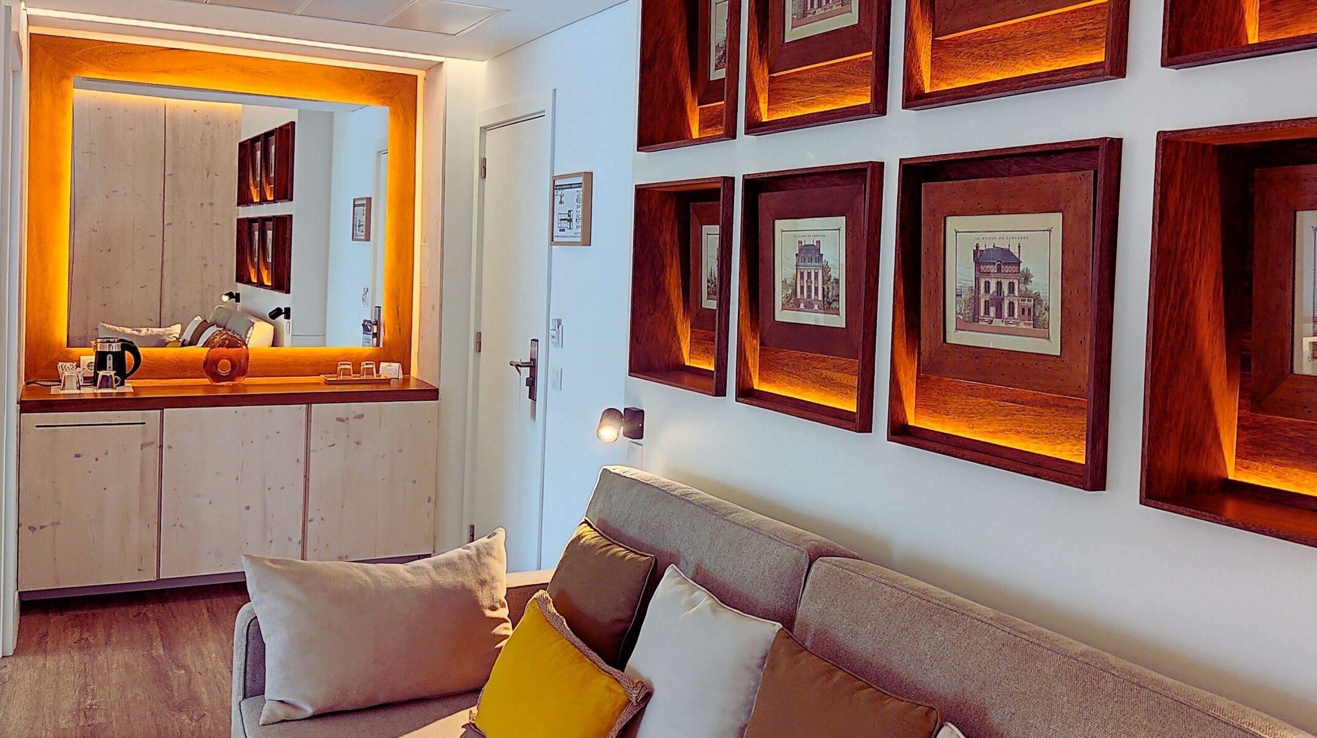 A modern hotel room with a wooden reception desk, a sofa with cushions, and framed pictures on the wall.