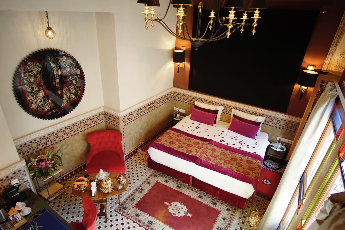 Una habitación con una cama, una silla roja, una mesita con un jarrón de flores y una araña.