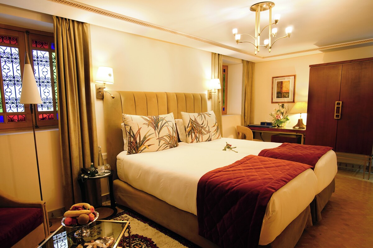 A hotel room with a large bed, a desk, a chair, a nightstand, and a window with curtains.