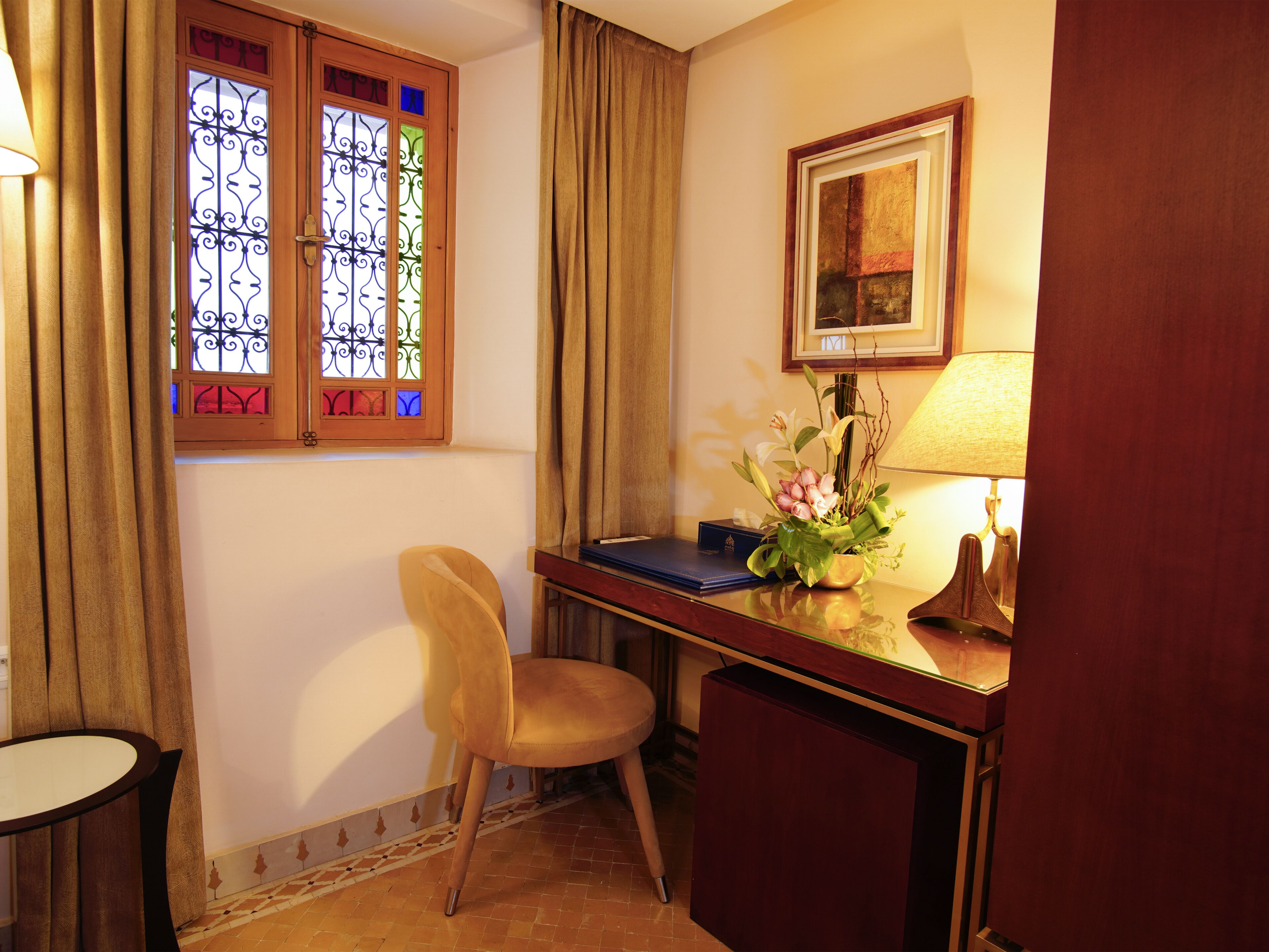 A room with a desk, chair, window with stained glass, lamp, and framed picture on the wall.