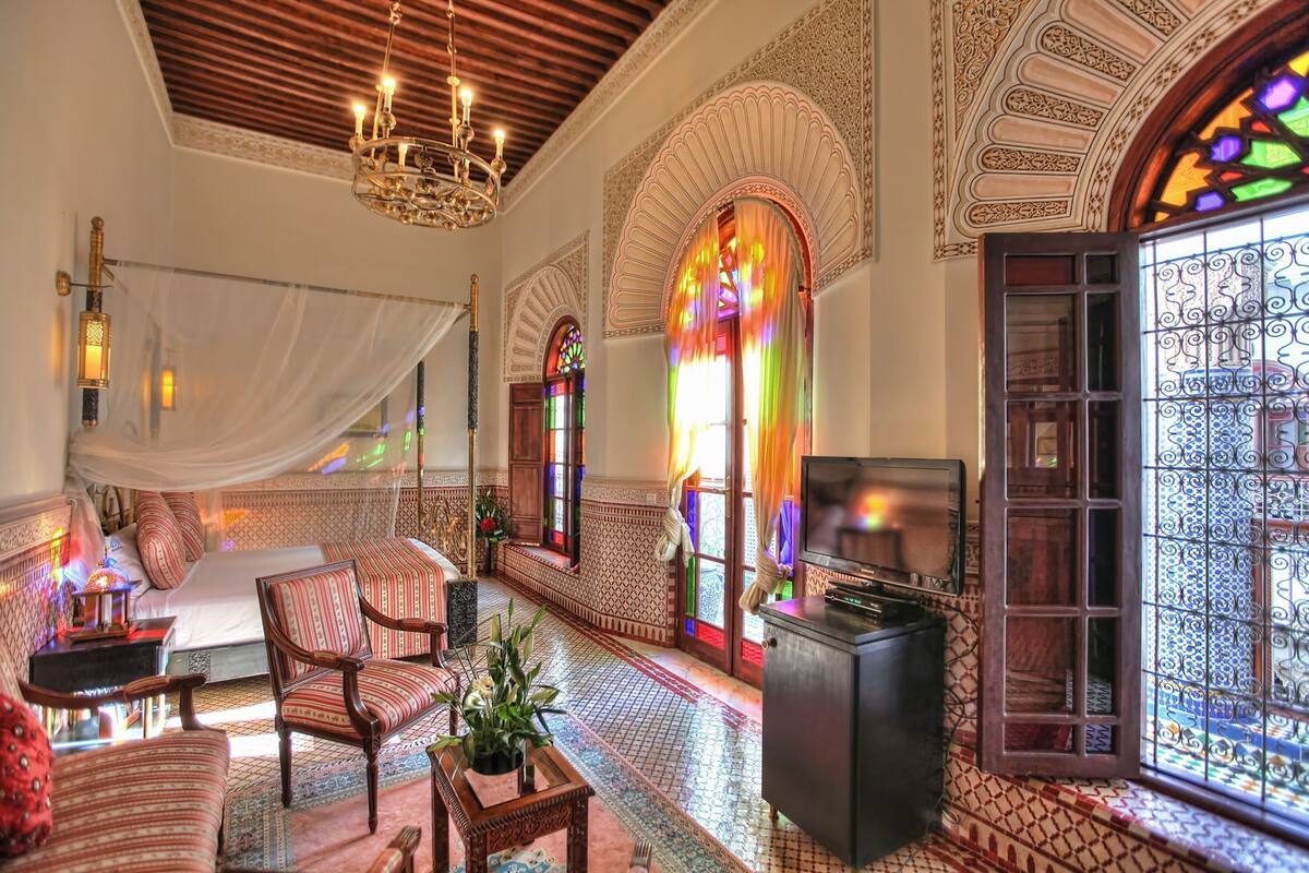 Una habitación con una cama, televisión, candelabro, sofá y un ventanal con vidrieras.