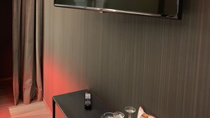 A hotel room with a flat-screen TV mounted on the wall, a small table with a remote control, glasses, and a snack package.