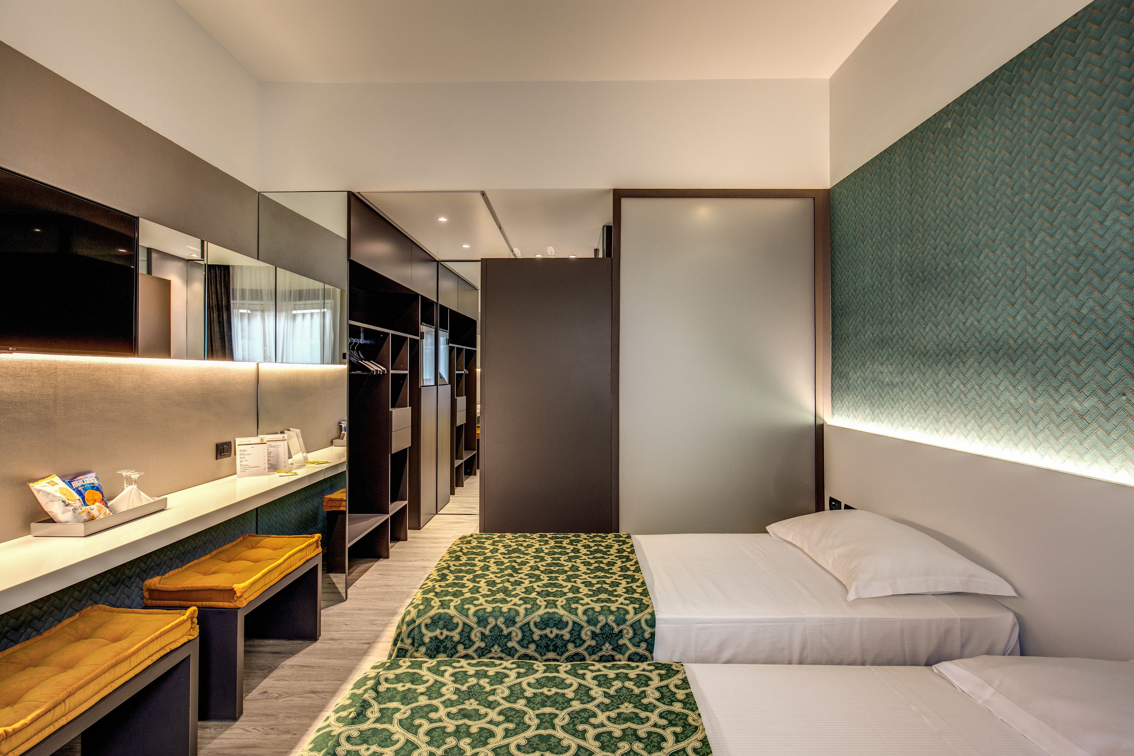 A modern hotel room with a large mirror, a bench, two beds with patterned bedding, and a wardrobe with a mirror.