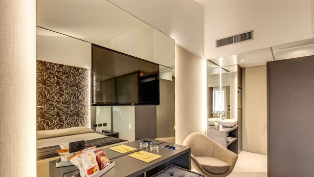 A modern hotel room with a large bed, a seating area with a table, a flat-screen TV, and a bathroom visible through a glass partition.