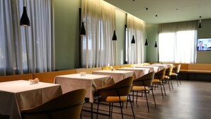 A restaurant with multiple tables and chairs, large windows with curtains, and hanging pendant lights.
