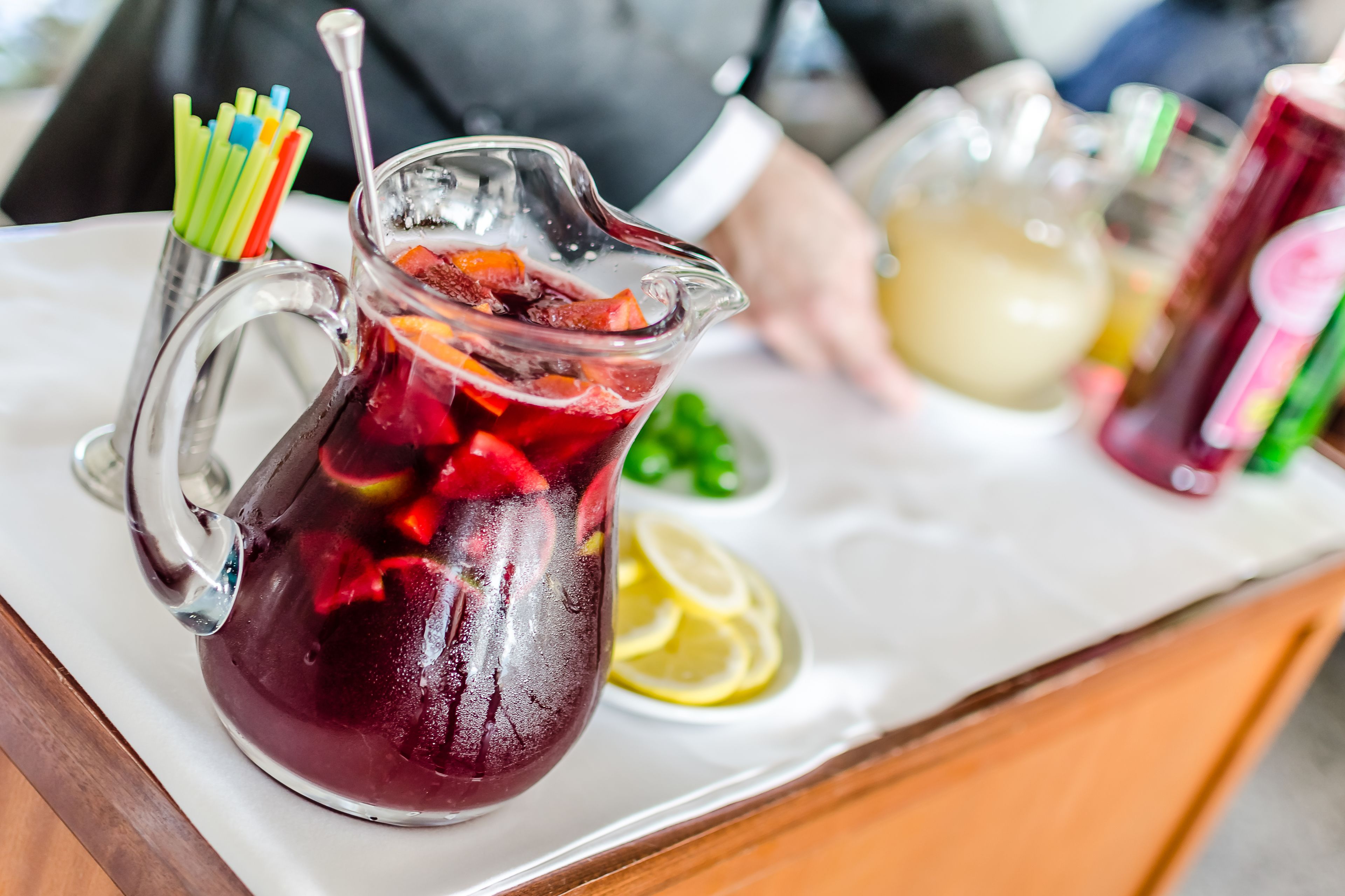 Un jarro de bebida helada con rodajas de fruta y una cuchara, un trozo de limón y un jarro de vidrio al fondo.