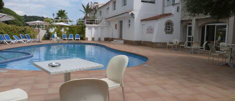 A pool area with a table and chairs, a building with a tiled roof, and a clear sky.