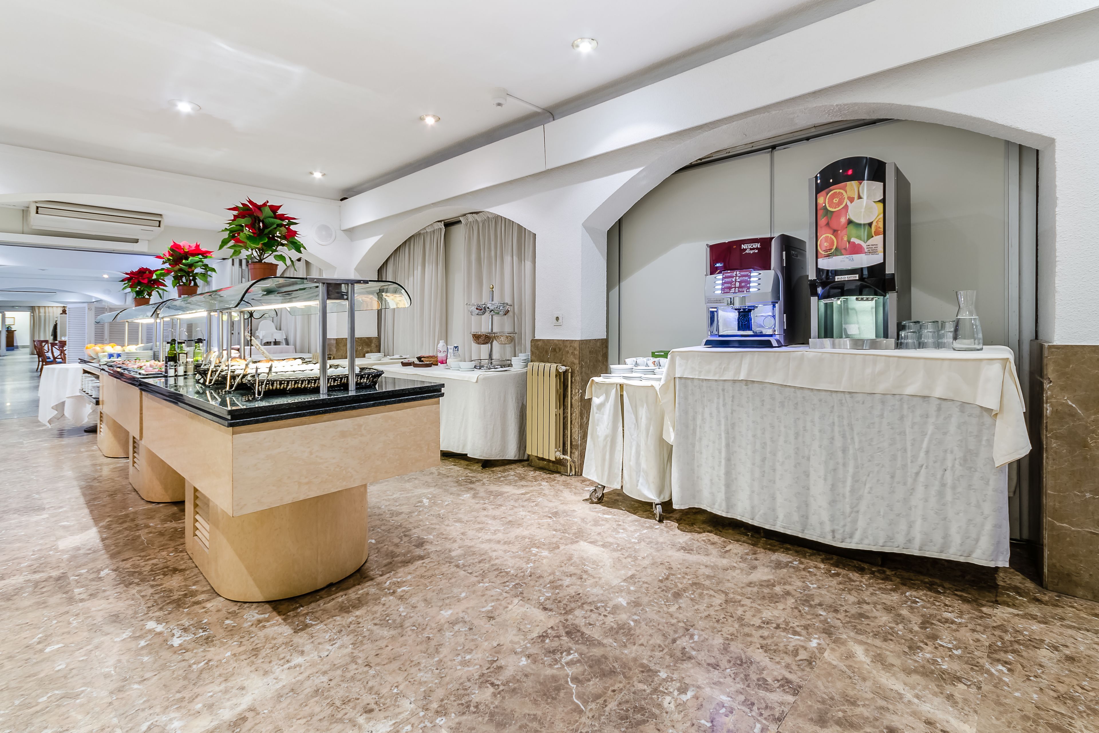 Eine moderne Hotelhalle mit Marmorboden, einem Buffetbereich mit einer Glasvitrine, einer Kaffeestation und Topfpflanzen.