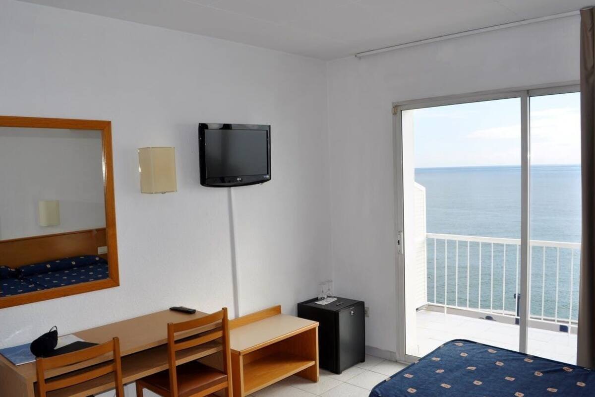 A hotel room with a bed, a desk with a chair, a television, a mirror, and a balcony with a view of the sea.