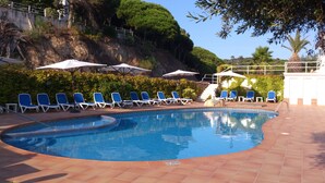 A swimming pool with blue water, surrounded by lounge chairs and umbrellas, with a rocky hillside in the background. - Hotel Gran Sol (Sant Pol De Mar)