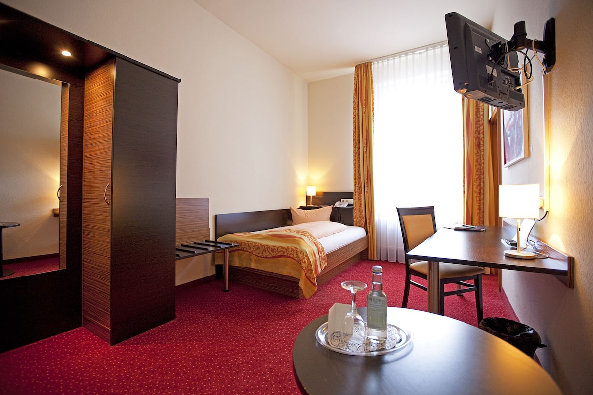 A hotel room with a bed, bedside table, lamp, chair, and a TV mounted on the wall.