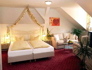 Un dormitorio con una cama doble, un rincón de descanso con sofá y sillones, una mesita con un florero y lámpara, y una planta grande.