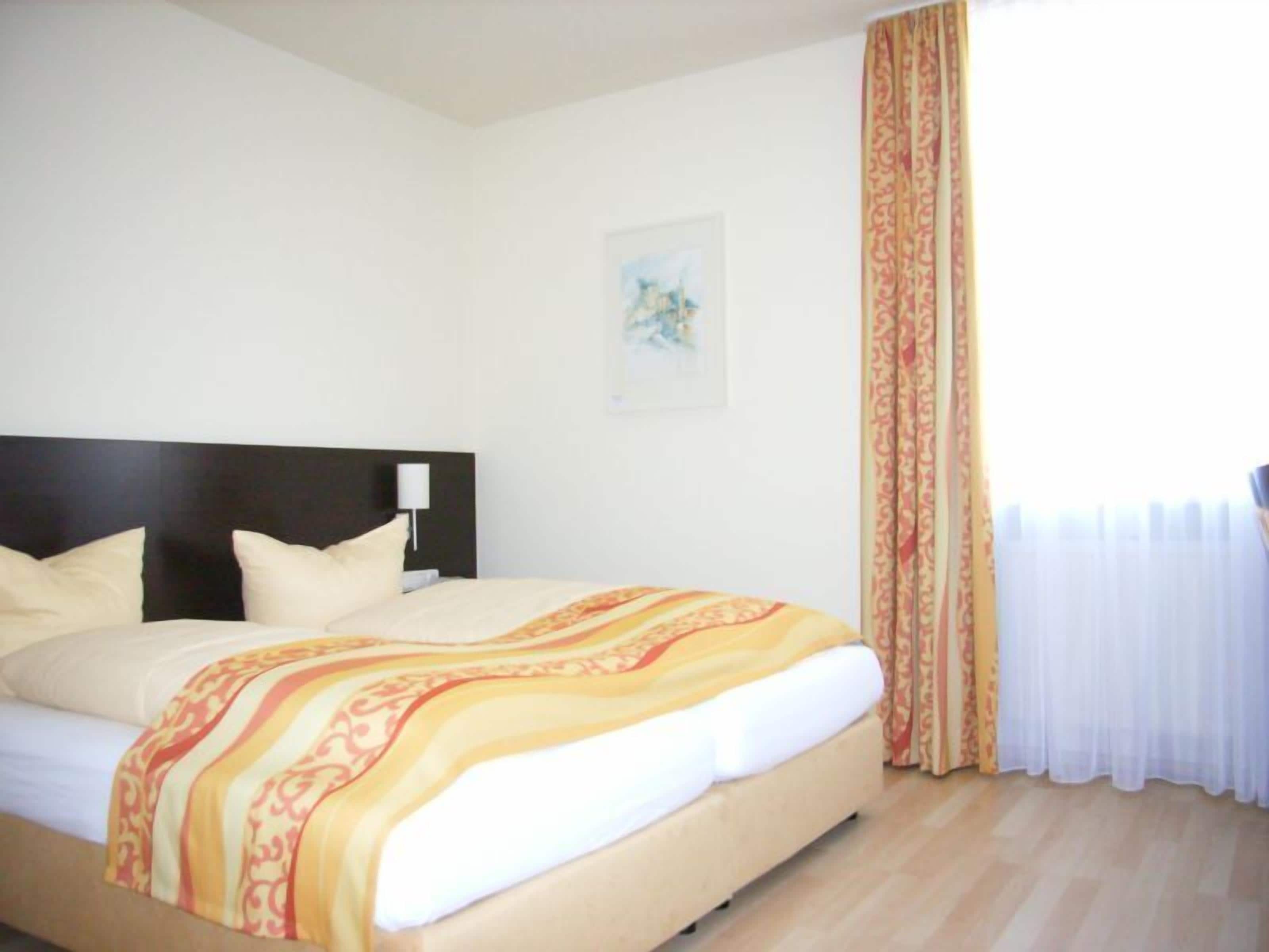 Una habitación de hotel con cama, cabecera, mesita de noche, lámpara, ventana con cortinas y un cuadro en la pared.