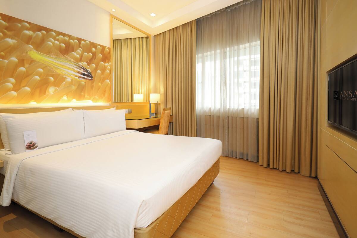 A hotel room with a large bed, a wooden headboard, a flat-screen TV, and a large window with curtains.