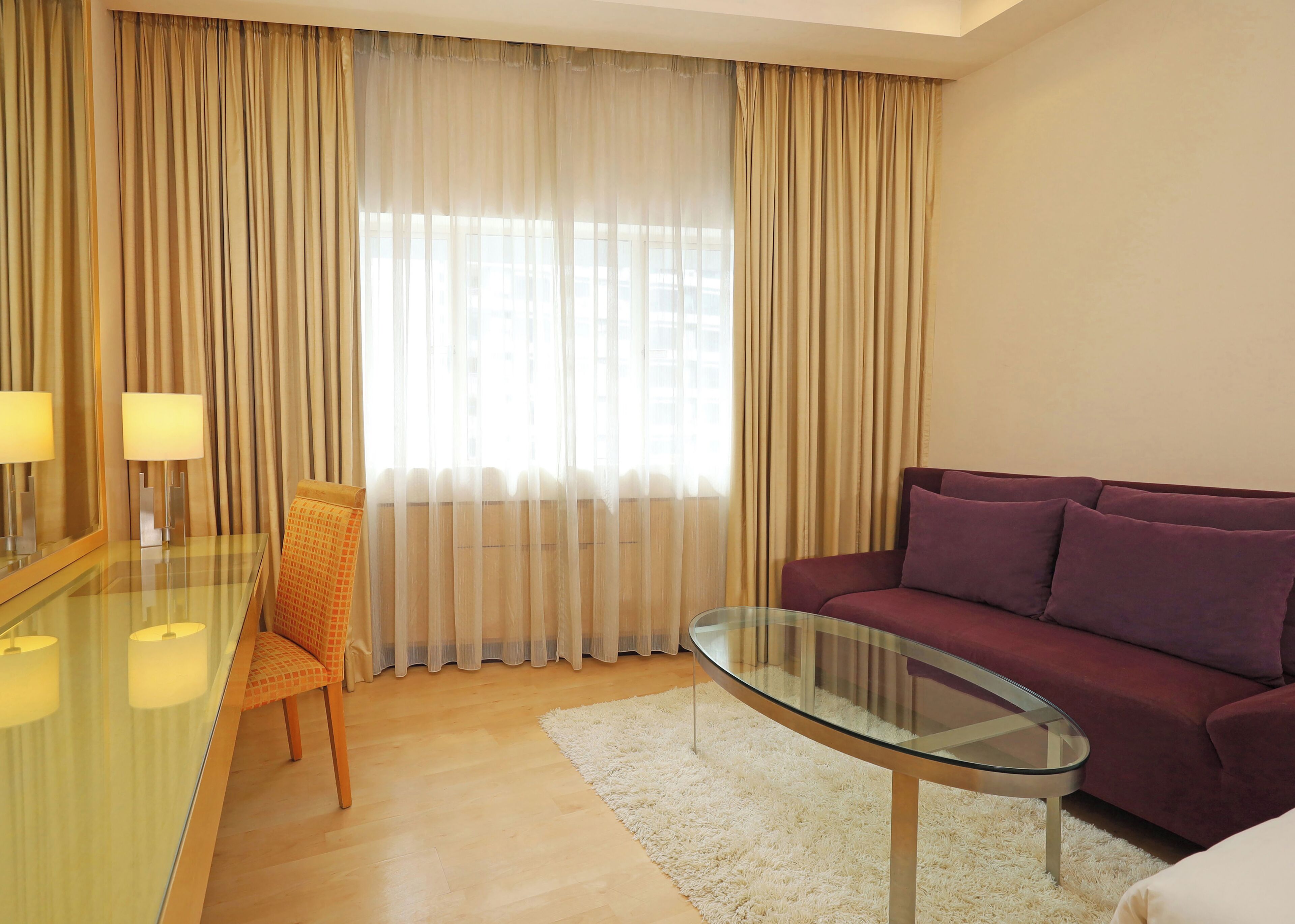 Una sala de estar moderna con una mesa de centro de vidrio, un sofá morado, una silla y un ventanal con cortinas translúcidas.