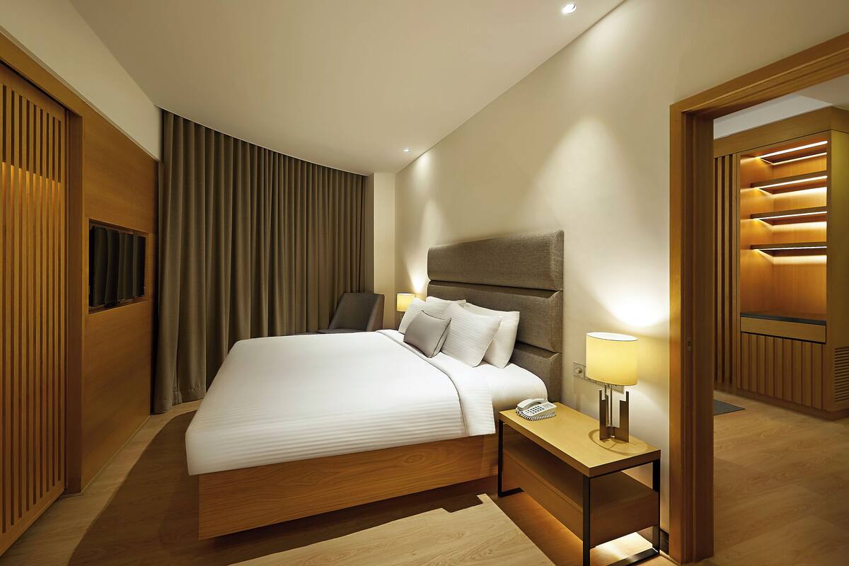 A modern hotel room with a large bed, a bedside table with a lamp, a wooden headboard, and a built-in shelf with lighting.