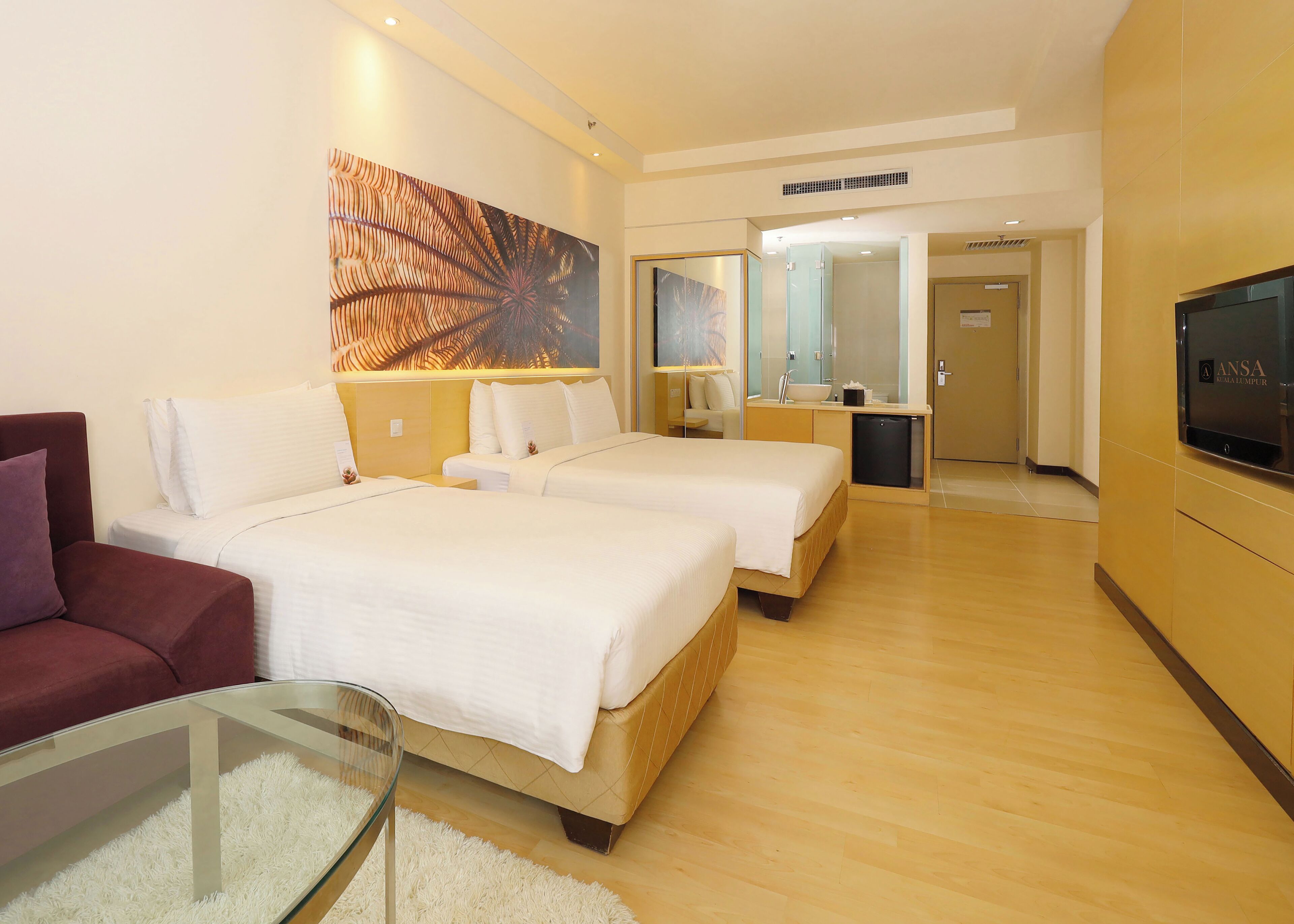 Habitación de hotel con dos camas, un sofá, un televisor, una obra de arte grande y un baño visible a través de una puerta abierta.