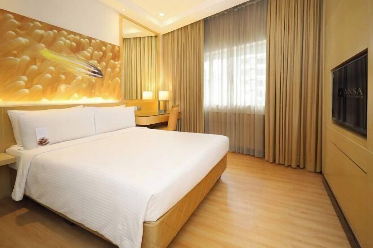 A hotel room with a large bed, a wall-mounted TV, a bedside table with a lamp, and a large window with curtains.