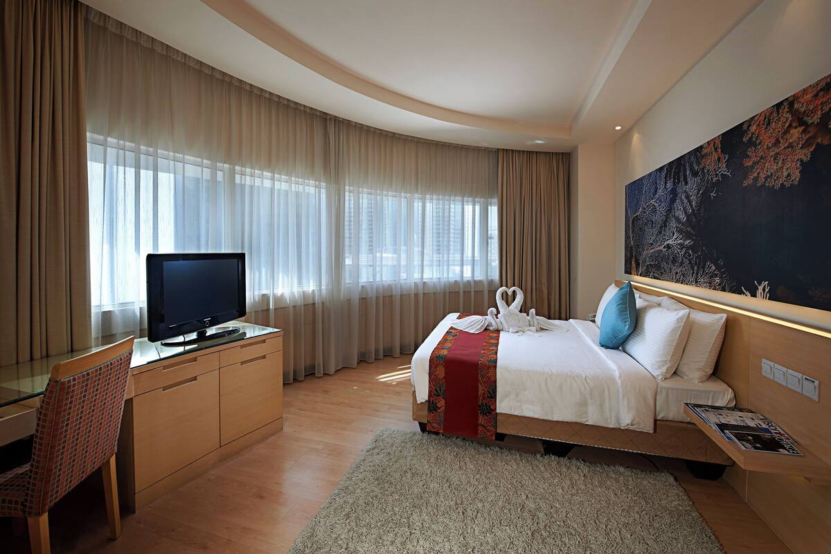 A modern hotel room with a flat-screen TV, a desk with a chair, a large bed with a red and white blanket, and a large painting on the wall.