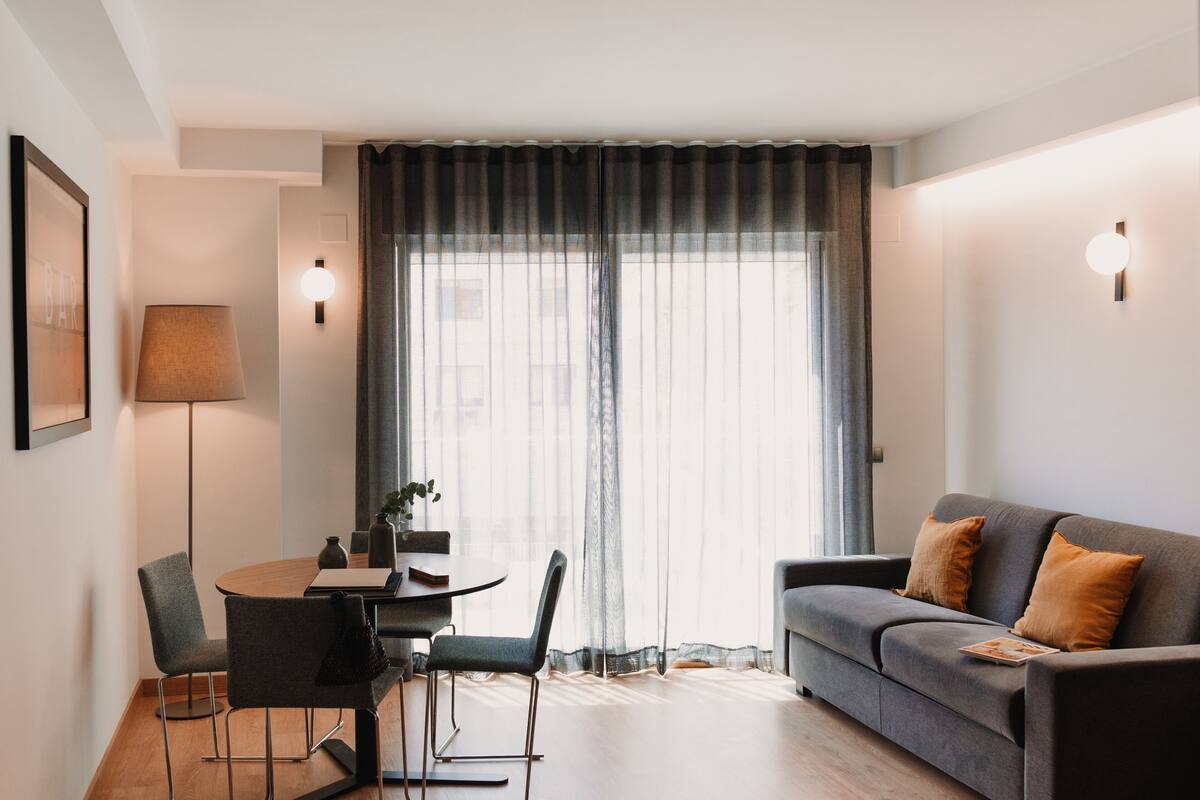 Ein modernes Wohnzimmer mit einer Couch, einem Esstisch mit Stühlen, einer Stehlampe und transparenten Vorhängen.