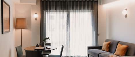 A modern living room with a dining area, featuring a sofa, chairs, a table, and a large window with curtains.