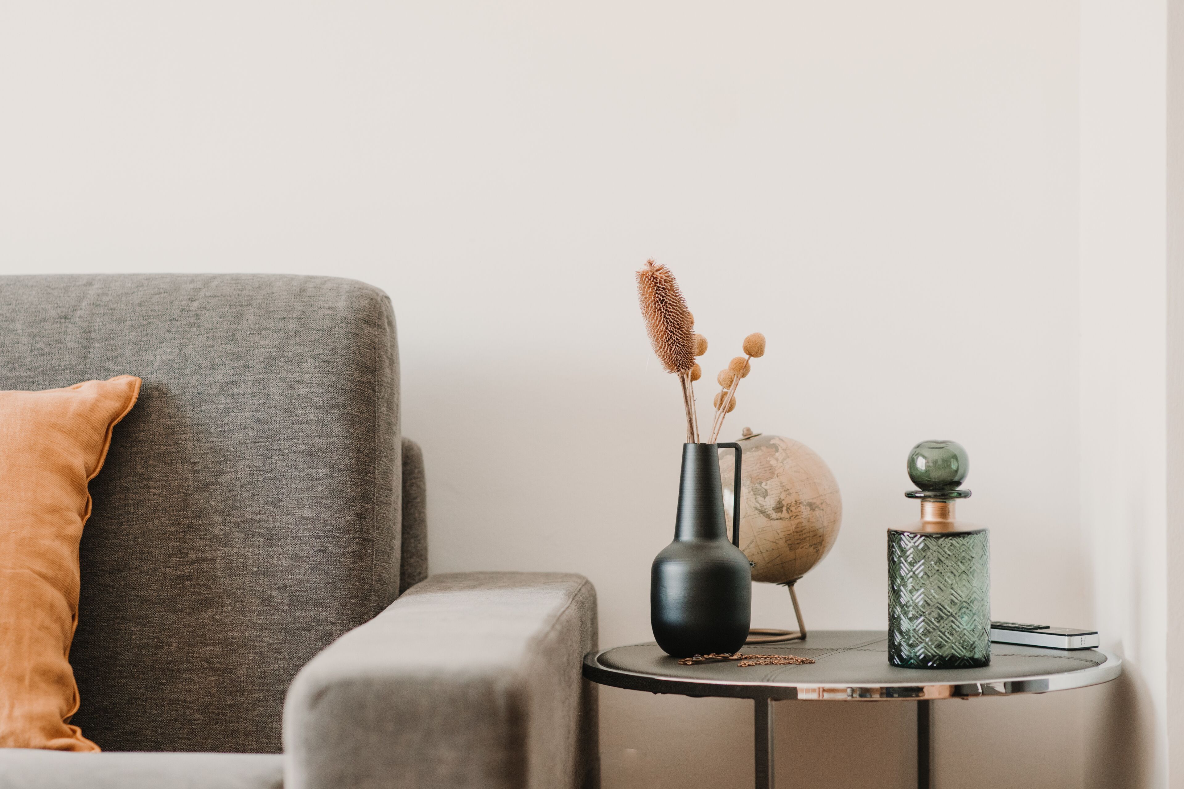 A grey sofa with a brown cushion, a small round table with a globe, a black vase with dried plants, and a green patterned vase.